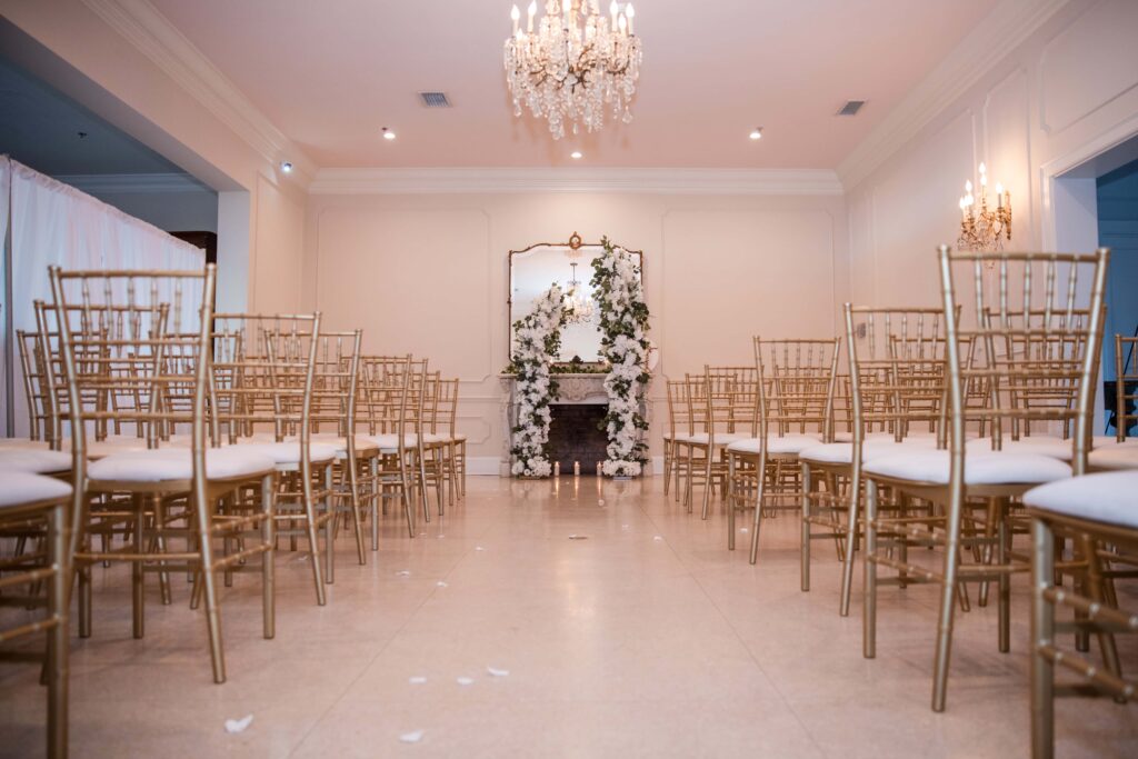 Indoor wedding ceremony setup at Azaleana Manor with gold chairs and white floral fireplace arch.