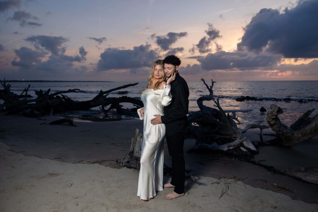 Elegant engagement portrait at golden hour with dramatic clouds. Bride-to-be in satin gown standing with fiancé during beach session.