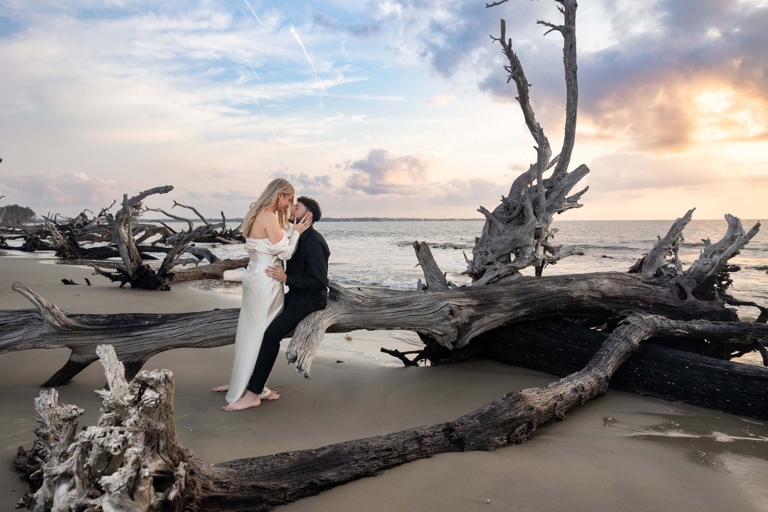 Bride-to-be in satin dress holding fiancé’s face. Sunrise engagement session at Driftwood Beach with couple sitting on sculpted driftwood by the water