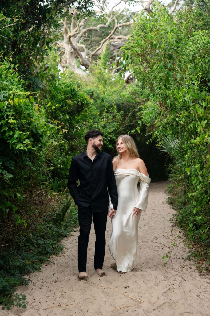 Engaged couple walking barefoot on a lush green path to Driftwood Beach, Jekyll Island during engagement session