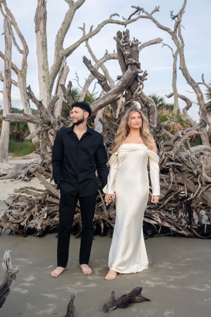 Engaged couple standing barefoot on Driftwood Beach in front of twisted driftwood trees during elegant engagement session